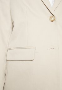 Béžový strukturovaný blazer s klopovým límcem, jedním knoflíkem, texturovanou ribbovanou tkaninou a kapsou na levé straně.