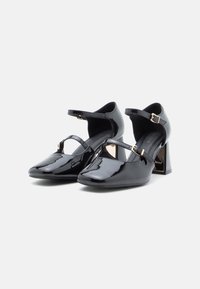 Black patent leather Mary Jane heels with square toes, ankle straps with gold buckles, and block heels on a white background.