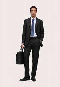 Schwarzer Anzug mit maßgeschneiderter Passform, weißes Hemd und blaue Krawatte. Trägt eine schwarze Ledertasche. Schwarze Schuhe, schlichte Textur, minimalistisches Design.