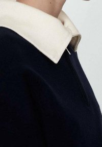 Pull-over bleu marine avec un col contrastant crème et une fermeture à glissière frontale mi-longue. Le tissu semble doux avec une finition texturée.