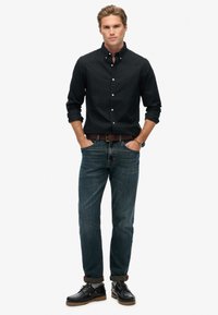 Sort skjorte med knapper, blå denim jeans og mørke sko. Bukserne rullet ved hælene. Brun bælte og ur synlige. Enkel, afslappet design.