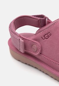 Rožinės zomšos sandalai su atitinkamu reguliuojamu diržu, tekstūruota pamušalu ir rudos gumos padu. Šone yra iškilni UGG prekės ženklo emblema.