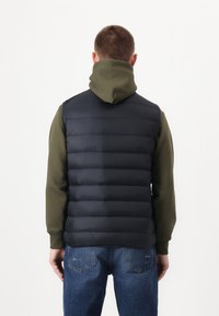 Navy puffer vest with horizontal quilted design, paired with an olive green hooded sweatshirt, worn with blue denim jeans.
