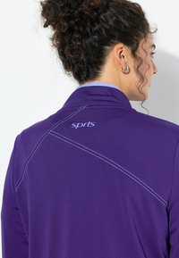 Fioletowa sportowa kurtka z białymi przeszyciami i jasnoniebieskim kołnierzem. Z tyłu małe logo "sprts". Gładka tekstura.