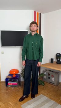 Uomo in camicia verde con bottoni e jeans scuri si trova in una stanza con una TV montata a parete, una lampada colorata, piante e una valigia con adesivi.