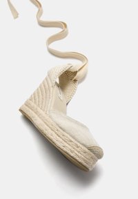 Beige espadrille kile sandal med tekstureret canvas overdel, vævet jute sål og lange stofbånd. Har en rund tå og en let hævet hæl.