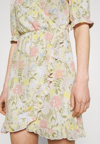 Blumen-Wickelkleid aus leichtem Stoff mit rüschengesäumtem Saum. Hat pastellfarbene Blumen in Pink, Gelb und Grün auf einem beigen Hintergrund. Weiche Textur.
