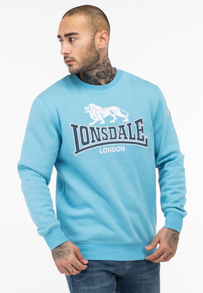 Világoskék pulóver, elöl logónyomattal, amelyen egy oroszlán és a "Lonsdale London" felirat található. Kerek nyakkivágással és bordázott mandzsettával tervezve.