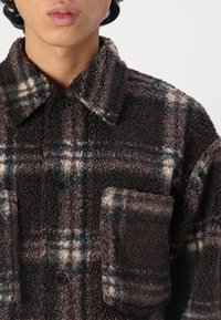 Veste en flanelle à carreaux marron à la texture douce, avec un col, un fermoir à boutons et une poche poitrine. Les motifs incluent des accents noirs et crème.