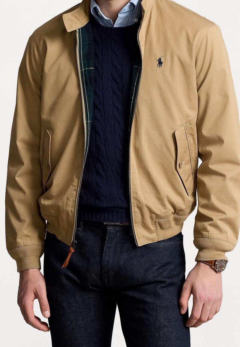 Beige bomberjack met gebreide manchetten en kraag, gecombineerd met een marineblauwe kabelgebreide trui en een lichtblauw overhemd, gedragen met een donkere spijkerbroek.