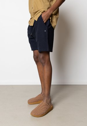 Persona che indossa pantaloncini blu navy, una maglietta beige e scarpe slip-on marrone intrecciate, in piedi con una mano in tasca su un pavimento chiaro.