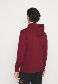 Sudadera burdeos con un acabado texturizado y capucha ajustable con cordón. Presenta puños y dobladillo acanalados, combinada con pantalones negros que exhiben un logo blanco.