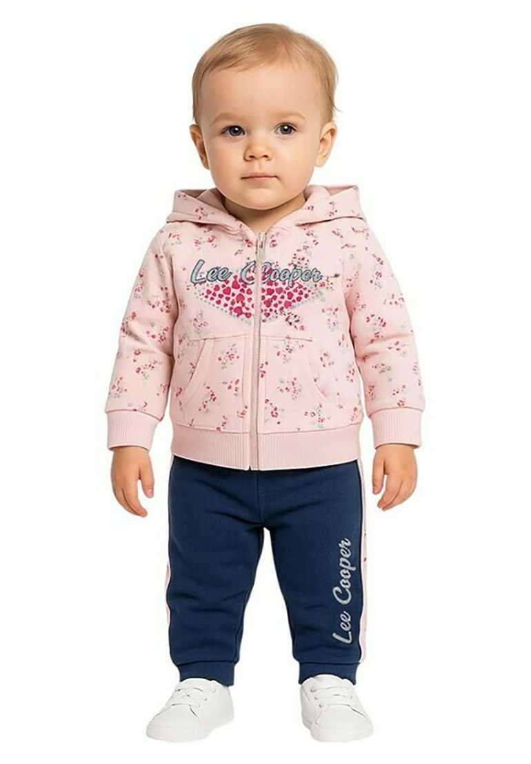 Sweat à capuche rose avec imprimé floral et fermeture éclair, associé à un pantalon de survêtement bleu marine avec des lettres blanches sur le côté. Baskets blanches.