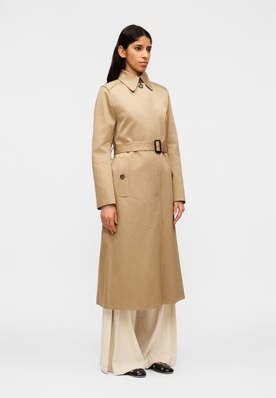 Γυναίκα που στέκεται, φορώντας ένα μακρύ μπεζ παλτό τρανς με ζώνη πάνω από φαρδιά κρεμ παντελόνια και μαύρα παπο ύτσια, σε απλό λευκό φόντο.