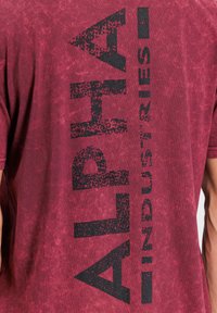 Červené, texturované bavlněné tričko s krátkými rukávy, na zádech s velkým, opotřebovaným černým potiskem se nápisem "ALPHA INDUSTRIES".