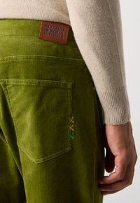 Grønne corduroypants har en tan lædermærke med præget branding og dekorative broderede accenter i brun, grøn og blå.