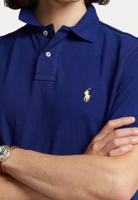 Personne portant un polo bleu avec un logo brodé jaune représentant un cheval et un cavalier, bras croisés, et une montre-bracelet avec un bracelet en cuir marron.