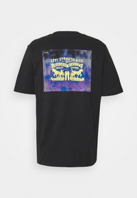 Černé bavlněné tričko s velkým fialovo-modrým tie-dye potiskem a žlutým logem Levi Strauss & Co. s nýtovým designem na zadní straně.
