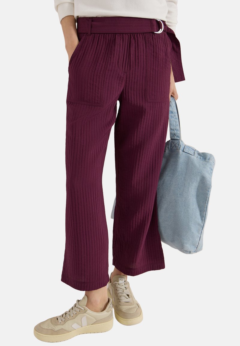 Femme portant un pantalon violet taille haute à larges jambes avec ceinture et baskets crème, portant un grand sac cabas bleu clair.