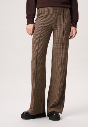 Vrouw die bruine wijde broek met pied-de-pouleprint en zwarte dikke plateauzolen draagt, staand tegen een effen lichte achtergrond.