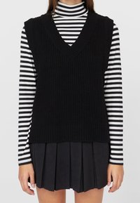 Veste noire en maille avec un col en V profond, portée sur un col roulé rayé noir et blanc et une jupe plissée noire. Tissu texturé.