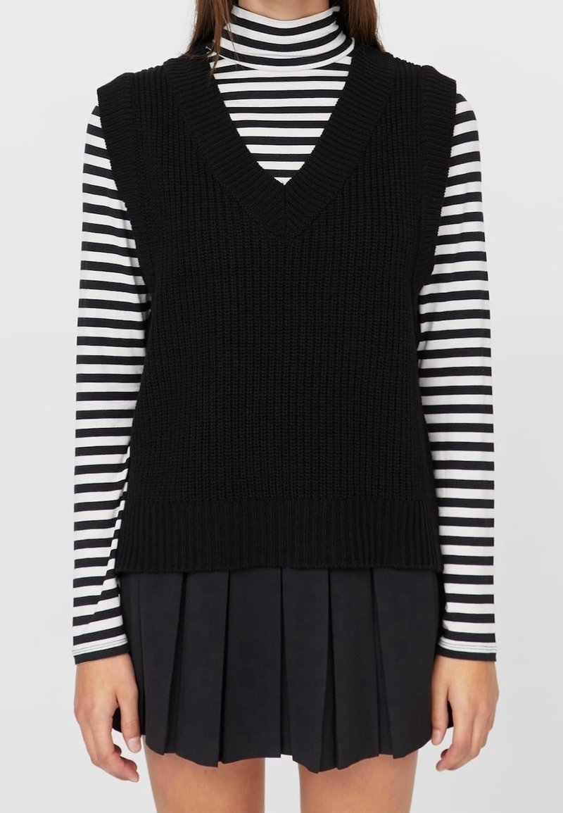 Veste noire en maille avec un col en V profond, portée sur un col roulé rayé noir et blanc et une jupe plissée noire. Tissu texturé.