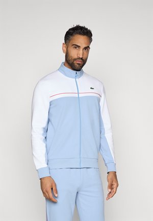 Ανοιχτό μπλε φούτερ με φερμουάρ και λευκά μανίκια, κόκκινη λωρίδα σε όλη την περιοχή του στήθους, ριμπ μανσέτες και λογότυπο Lacoste στην αριστερή πλευρά.