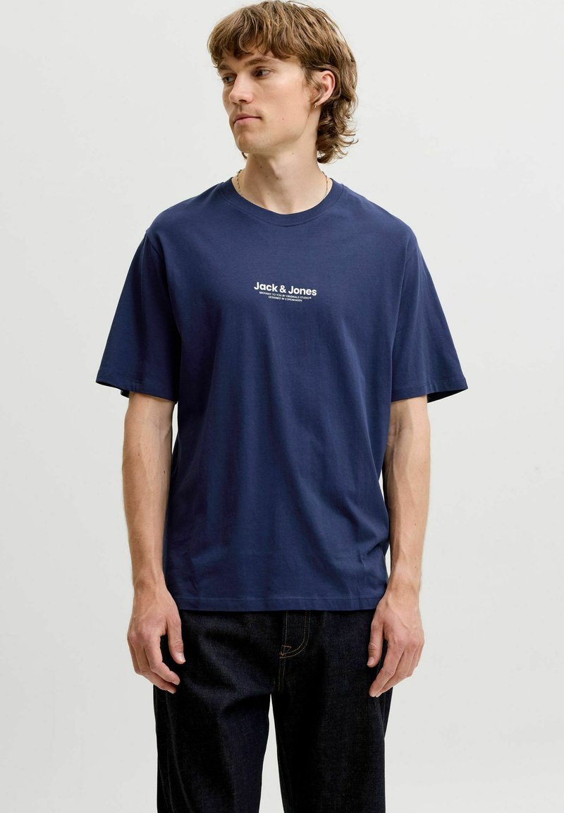 Jeune homme portant un t-shirt bleu marine Jack & Jones et un pantalon noir, debout devant un fond uni gris clair.