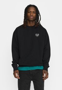 Zwarte sweatshirt met ronde hals en lange mouwen, met de witte tekst "OFF TO A GOOD START" op de borst. Gedragen over een turquoise shirt.