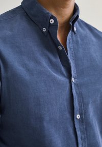 Navy blue, napakan hangat, na-button-up na shirt na gawa sa makinis na tela na may pointed collar. May puting mga butones at kaunting teksturadong finish.
