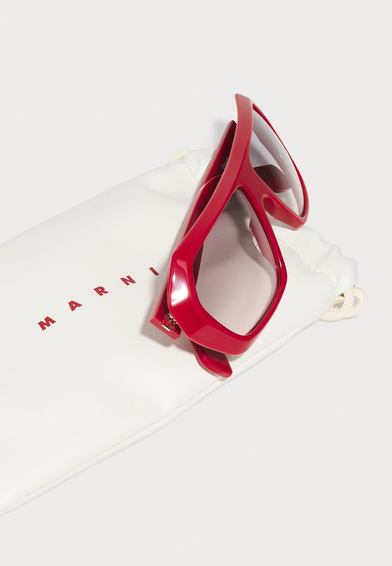 Rote übergroße Sonnenbrille mit dicker Fassung, die auf einem weißen, weichen Beutel mit roter Schrift "MARNI" liegt. Glatte Textur, modernes Design.