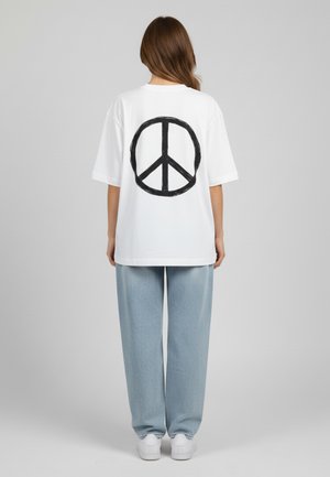 Hvid oversized t-shirt med et sort fredssymbol grafisk motiv på bagsiden. Sat sammen med lyseblå vide bukser og hvide sneakers.