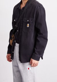 Svart denimskjorta med två framfickor och tryckknappar, bärs över en svart topp. Grå mjukisbyxor med "Champion"-logotyp vid höften.