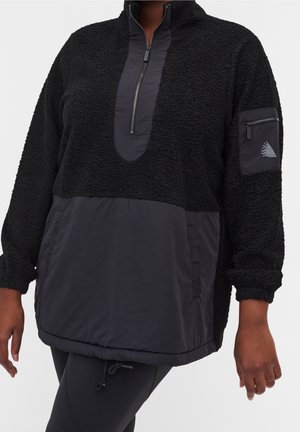 Pull-over en polaire noire avec demi-fermeture éclair, poches frontales et latérales, poignets élastiques, et une poche zippée sur la manche gauche.
