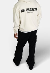 Felpa crema con la scritta "NO REGRETS" in grassetto nero sul retro, abbinata a pantaloni cargo neri larghi, dotati di tasche e un tessuto testurizzato.