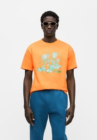 Πορτοκαλί βαμβακερό μπλουζάκι με πράσινο γραφικό σχέδιο που απεικονίζει χαρακτήρες και το λογότυπο της Nike. Φορεμένο με μπλε φόρμα και γυαλιά ηλίου.