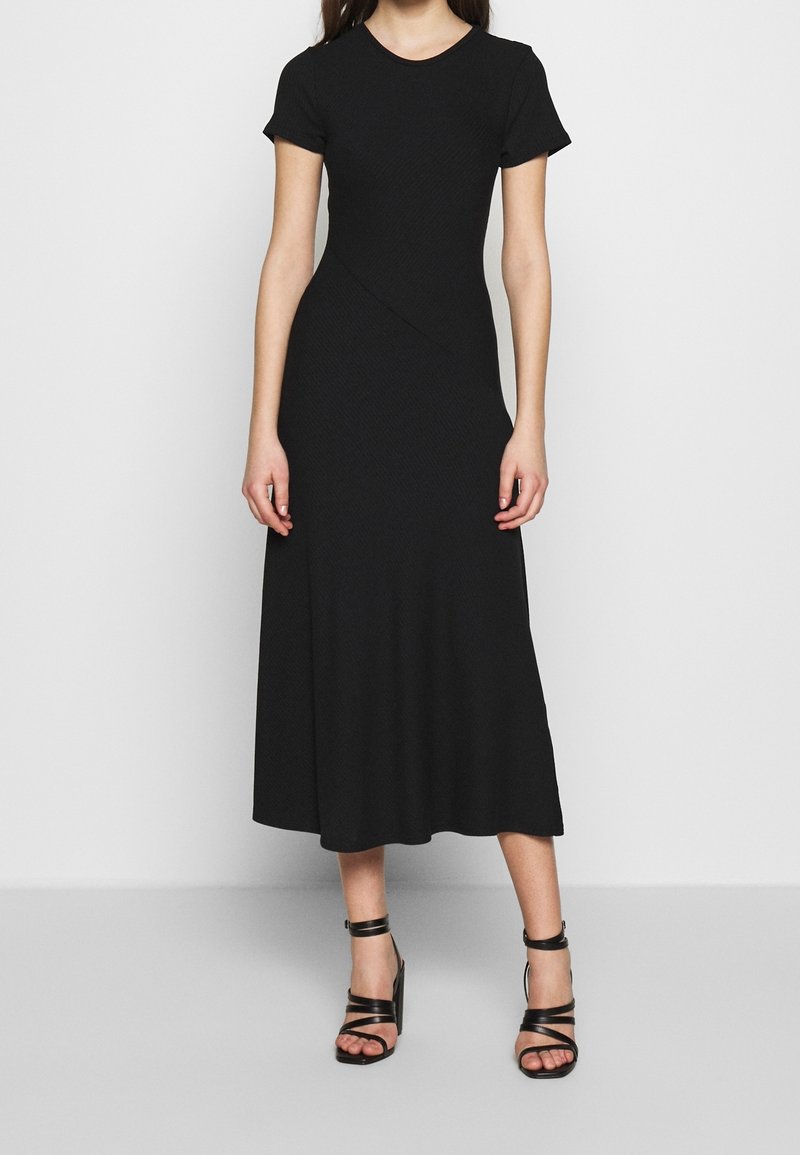Femme portant une robe midi noire cintrée à manches courtes avec une légère évasement, associée à des sandales noires à talons hauts et à lanières.