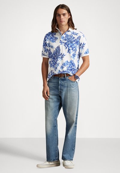 Πόλο μπλούζα με κοντά μανίκια και μπλε λουλουδάτο σχέδιο σε λευκό φόντο, συνδυασμένη με ξεβαμμένο μπλε τζιν με ίσια γραμμή και λευκά αθλητικά παπούτσια.