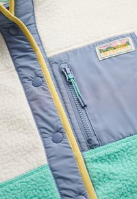 Close-up van een color-block fleecejack met blauwe drukknopen, een ritszak en een Superdry merklabel op licht- en muntgroene stof.