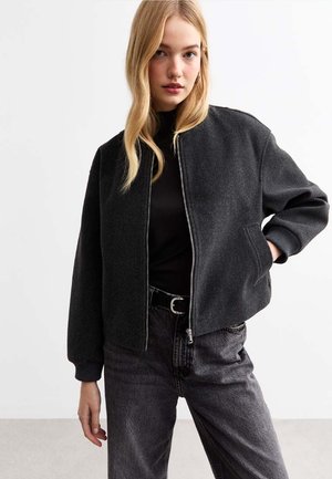 Chaqueta bomber de color gris oscuro con cierre de cremallera en el frente, puños acanalados y dobladillo elástico. Combinada con una camiseta negra y jeans grises, aparece en un entorno minimalista.