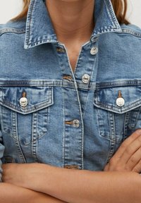 Vaaleansininen farkkutakki, jossa on terävä kaulus, hopeanväriset painonapit ja kaksi rintataskua. Kangas on teksturoitu ja siinä on hillitty haalistuma.