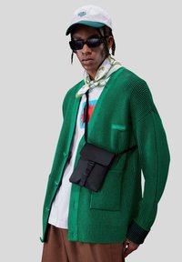 Grøn, ribbet cardigan med kontrasterende knapper, lagd over en hvid grafisk T-shirt. Sort crossbody taske, solbriller og mønstret tørklæde.