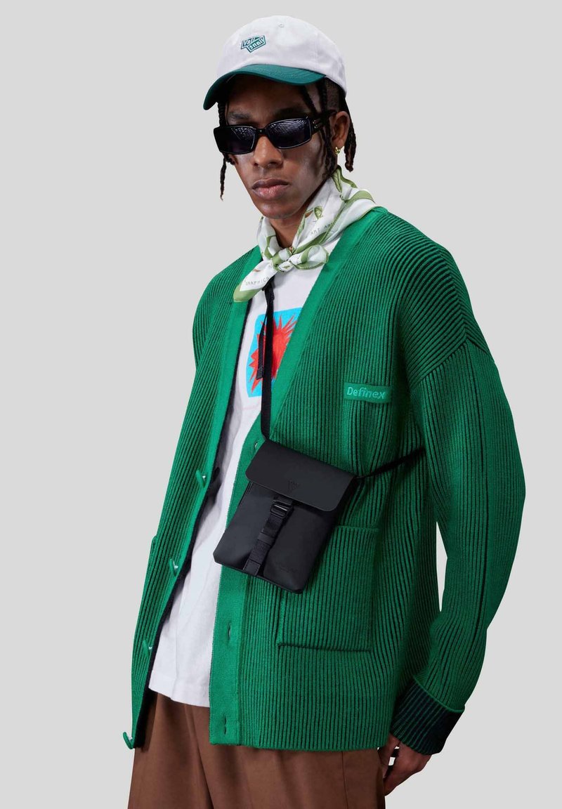 Grøn, ribbet cardigan med kontrasterende knapper, lagd over en hvid grafisk T-shirt. Sort crossbody taske, solbriller og mønstret tørklæde.