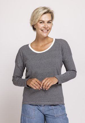 Smiling woman with short blonde hair wearing a black and white striped long-sleeve shirt and blue jeans against a plain background.