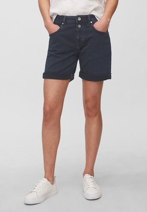 Person iført mørkeblå shorts med opkrogede kanter, hvide afslappede sneakers og en hvid skjorte, stående med hænderne i lommerne mod en ensfarvet baggrund.