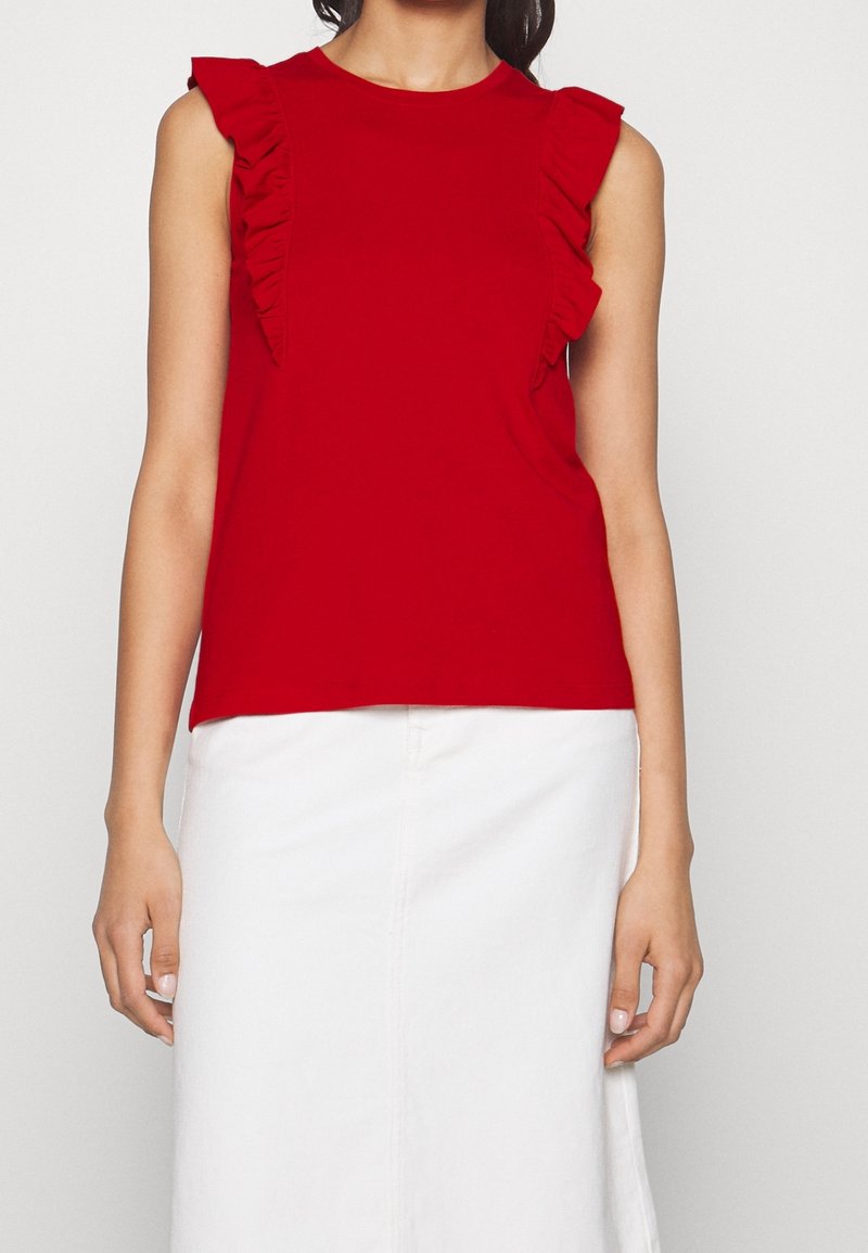 Červený top bez rukávů s volánovými akcenty na ramenou, vyrobený z měkké látky; v kombinaci s bílou sukní, vyzařující ležérní, moderní styl.