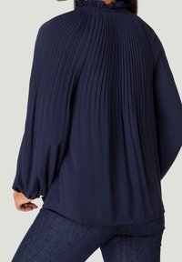 Blouse plissée bleu marine avec un col à volants et des manches longues ; confectionnée dans un tissu léger, avec une coupe décontractée.