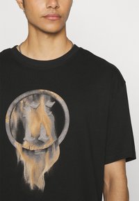 T-shirt en coton noir avec un graphique circulaire usé, comprenant une lettre stylisée en gris et or, créant un effet texturé et superposé.