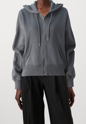 Persoon draagt een grijze capuchontrui met rits, voorzakken en trekkoorden, gecombineerd met zwarte wijde broek met krijtstrepen, staand tegen een effen achtergrond.