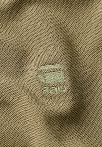 Il tessuto verde khaki presenta una trama testurizzata con un logo ricamato di colore verde chiaro che raffigura un'immagine stilizzata e il testo "RAW" sottostante.